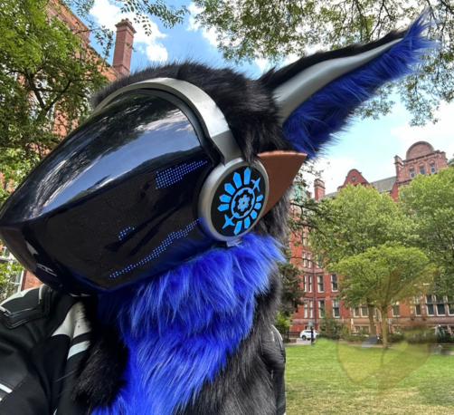 Alt: A black and blue furred protogen fursuit with blue LED face. Black and blue furred long ears with metallic silver strip along the length of the ear. Fursuit head has silver and bronze painted plastics and illuminated side panels. Photo is taken at a 3/4 right selfie angle. Background is trees, blue sky and a grassy park surrounded by red brick victorian buildings.