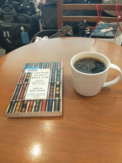 Photo is of a brown wooden table in the Starbucks. On the table is a white mug of black coffee. To the left is the paperback library book with an image of many old guilded books each shelved between white metal cells like a prison cell