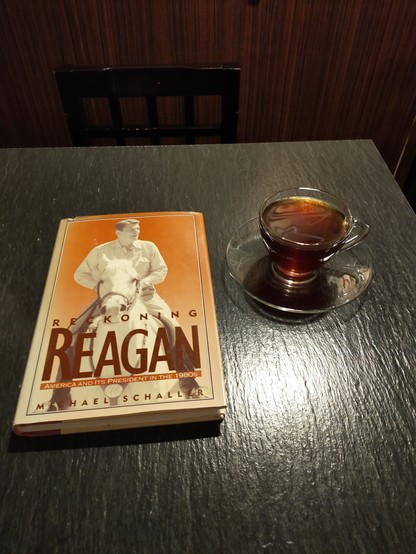 On a black wooden table is the orange hardcover book with cover of white man Ronald Reagan on a white horse. To the right is a clear mug & saucer of black coffee